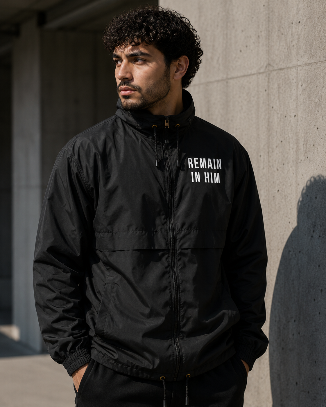 Man wearing a black jacket with 'REMAIN IN HIM' text against a concrete wall.
