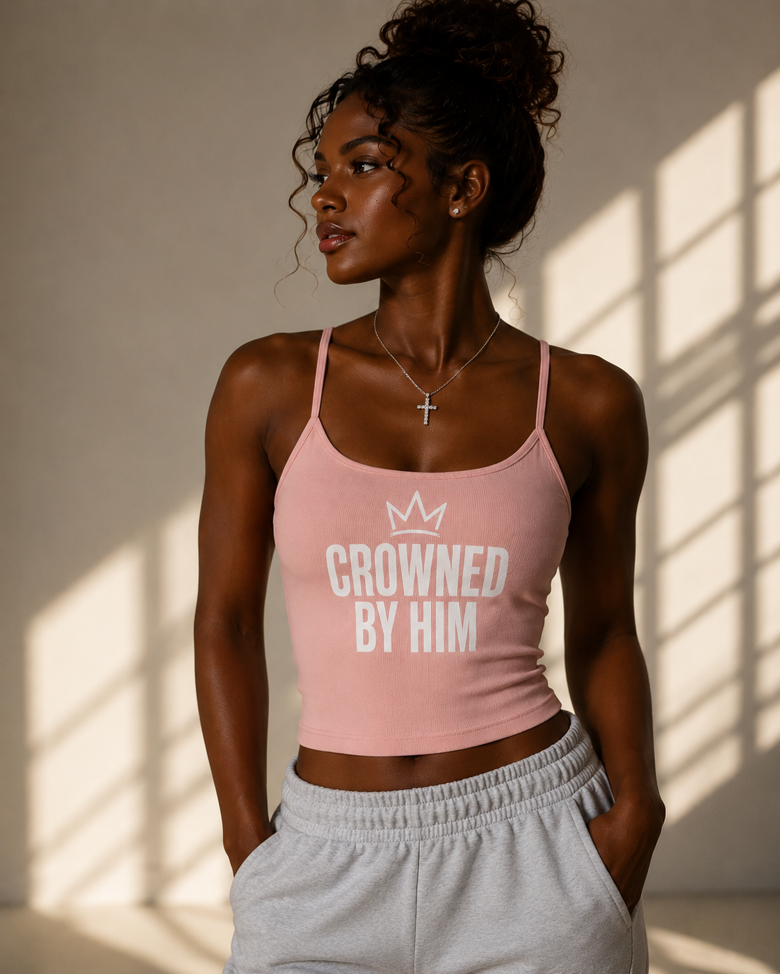 Woman wearing a pink tank top with 'Crowned by Him' text, standing in a softly lit room.