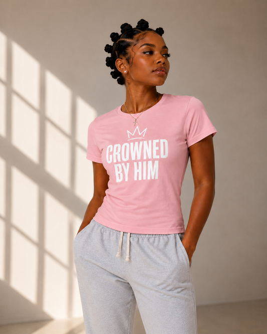 Woman wearing a pink t-shirt with 'Crowned by Him' text, standing in a sunlit room.