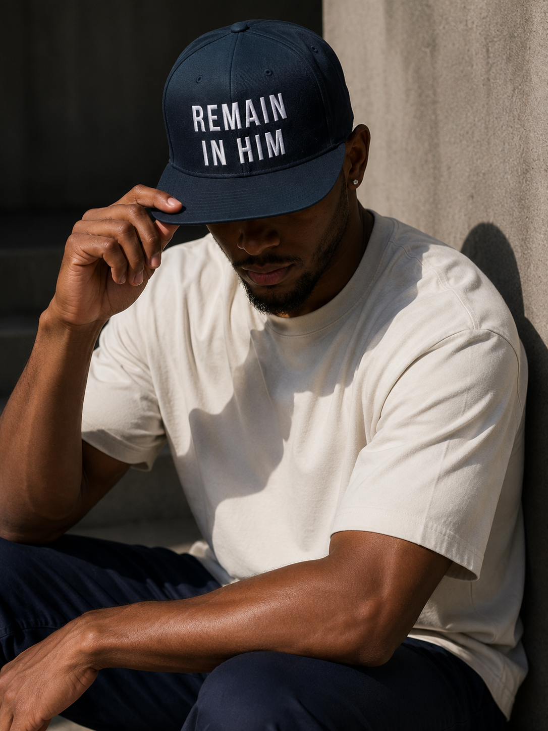Person wearing a navy cap with 'REMAIN IN HIM' text, sitting against a neutral background