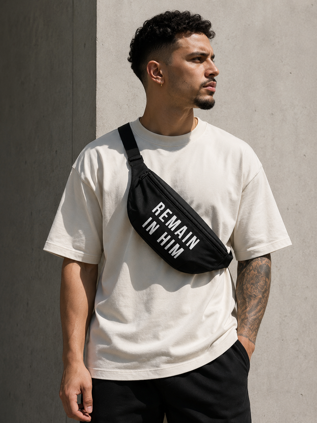Man wearing a white t-shirt and black waist bag with 'REMAIN IN HIM' text against a gray wall.