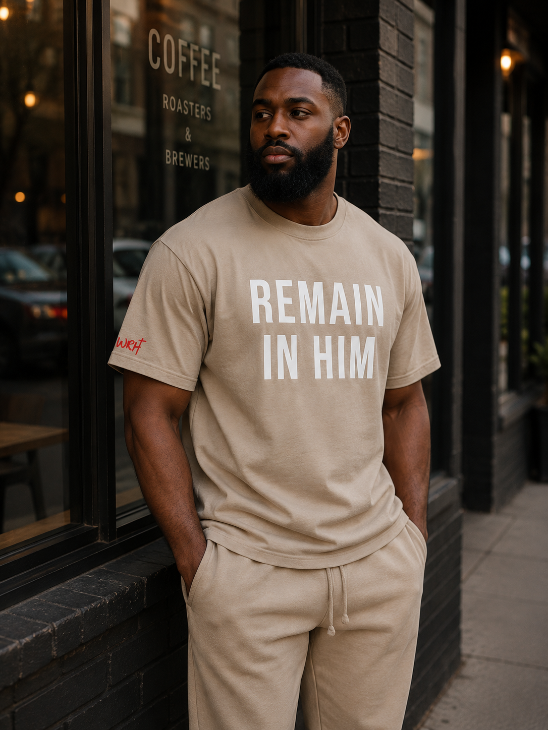 Man wearing a beige t-shirt with 'REMAIN IN HIM' text, standing outside a coffee shop.