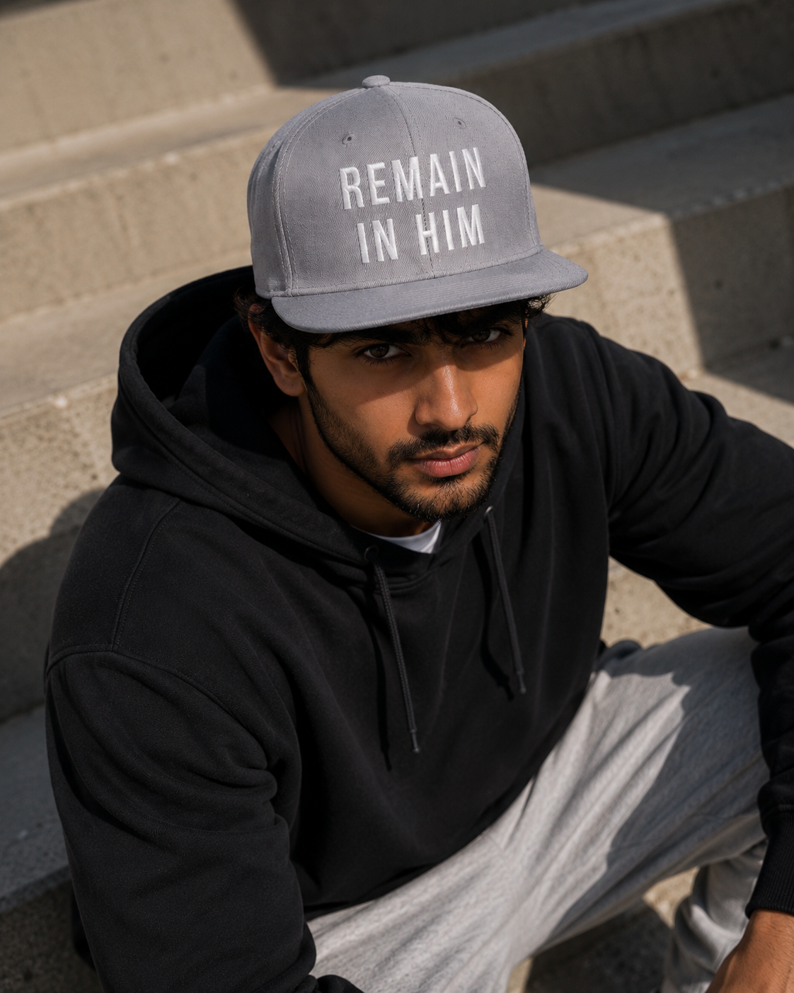 Man wearing a black hoodie and gray cap with 'REMAIN IN HIM' text, sitting on steps.