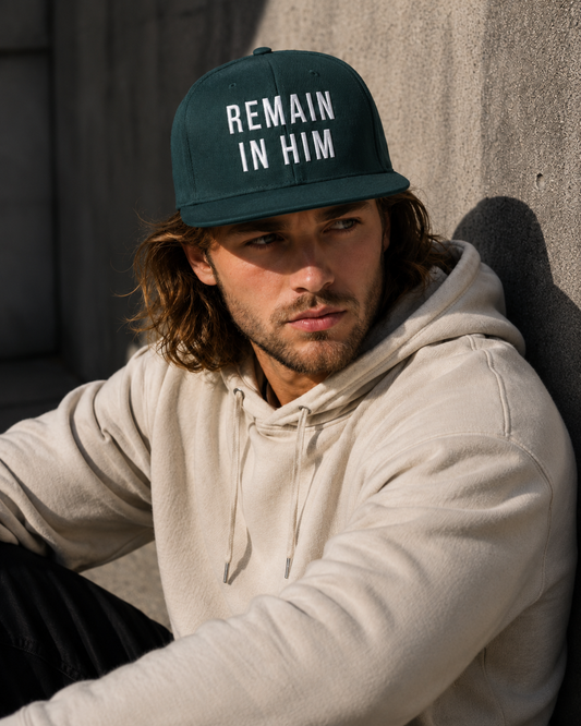 Person wearing a cap with 'REMAIN IN HIM' text, sitting against a concrete wall.